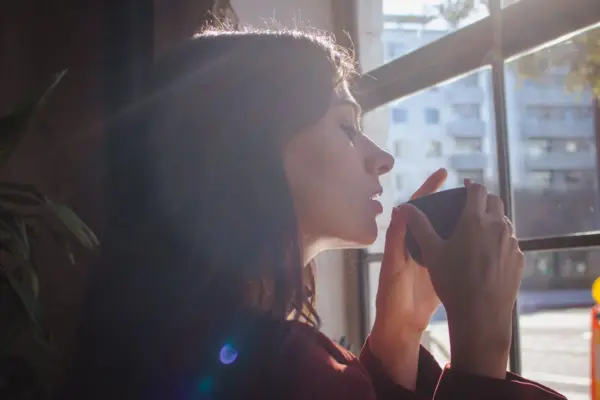 Mujer tomando una bebida caliente junto a la ventana, con calma